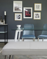 Modern dining room furniture featuring a sleek black glass-top table with white legs, accompanied by three blue upholstered dining chairs with slim metal legs. In the foreground, a contemporary light grey fabric bench with clean lines and subtle stitching details.