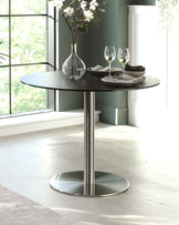 A modern round table with a sleek black top and stainless steel base, adorned with a vase, glasses, and dinnerware.