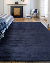 A contemporary room featuring a large, textured navy blue area rug on a rustic wooden floor. In the corner sits a modern white chair with black legs, accented by a navy blue throw casually draped over one side. The space is complemented by a classic white marble fireplace and natural light from a window with wooden shutters.