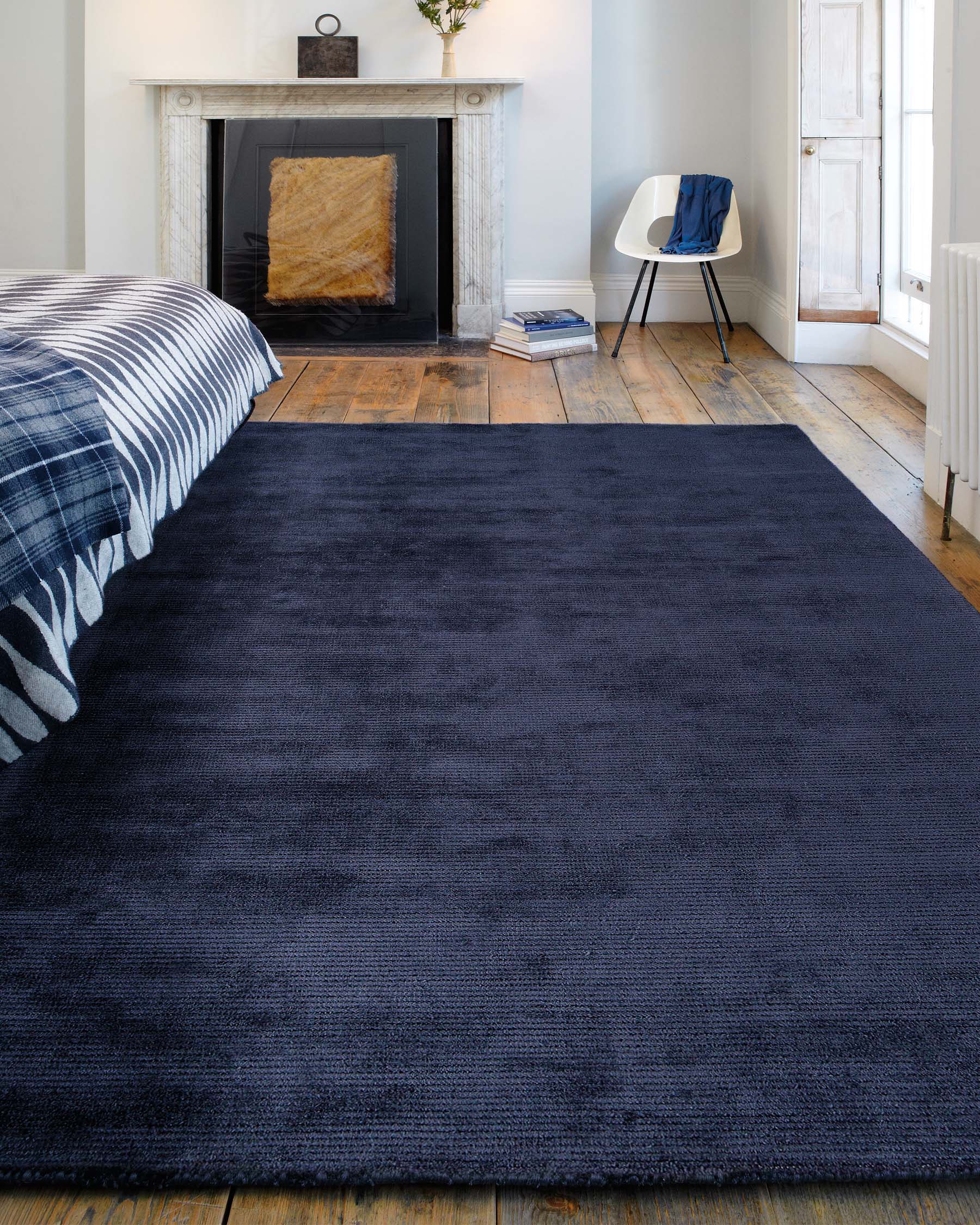 A contemporary room featuring a large, textured navy blue area rug on a rustic wooden floor. In the corner sits a modern white chair with black legs, accented by a navy blue throw casually draped over one side. The space is complemented by a classic white marble fireplace and natural light from a window with wooden shutters.