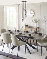 Modern dining room set with a rectangular grey table with black angular legs accompanied by six cream upholstered chairs with black metal legs, centred on a two-tone area rug. A sleek black console table with stacked shelves is placed against the wall, adorned with decorative items and a white vase with greenery.