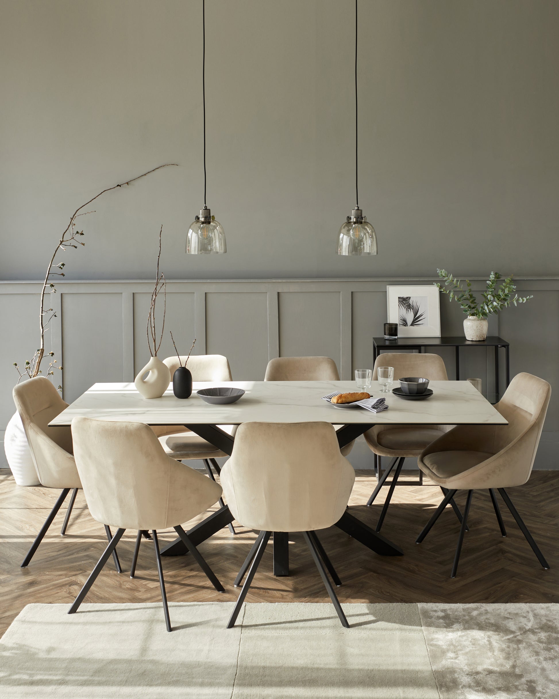 Modern dining room featuring a rectangular table with a light wood finish and black angular legs, surrounded by six beige upholstered chairs with a sleek curved design and black metal legs. The set is complemented by two clear glass pendant lights above the table, on a backdrop of a grey wall with wainscoting details. There is also a black side table with a small selection of decorative items and potted plants, enhancing the contemporary aesthetic.