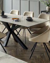 Modern dining set featuring a rectangular marble-top table with a distinctive geometric black metallic base. Paired with it are plush upholstered beige dining chairs with a sleek curved design and black metal legs. The set is presented in a contemporary room with wood flooring and grey panel walls.