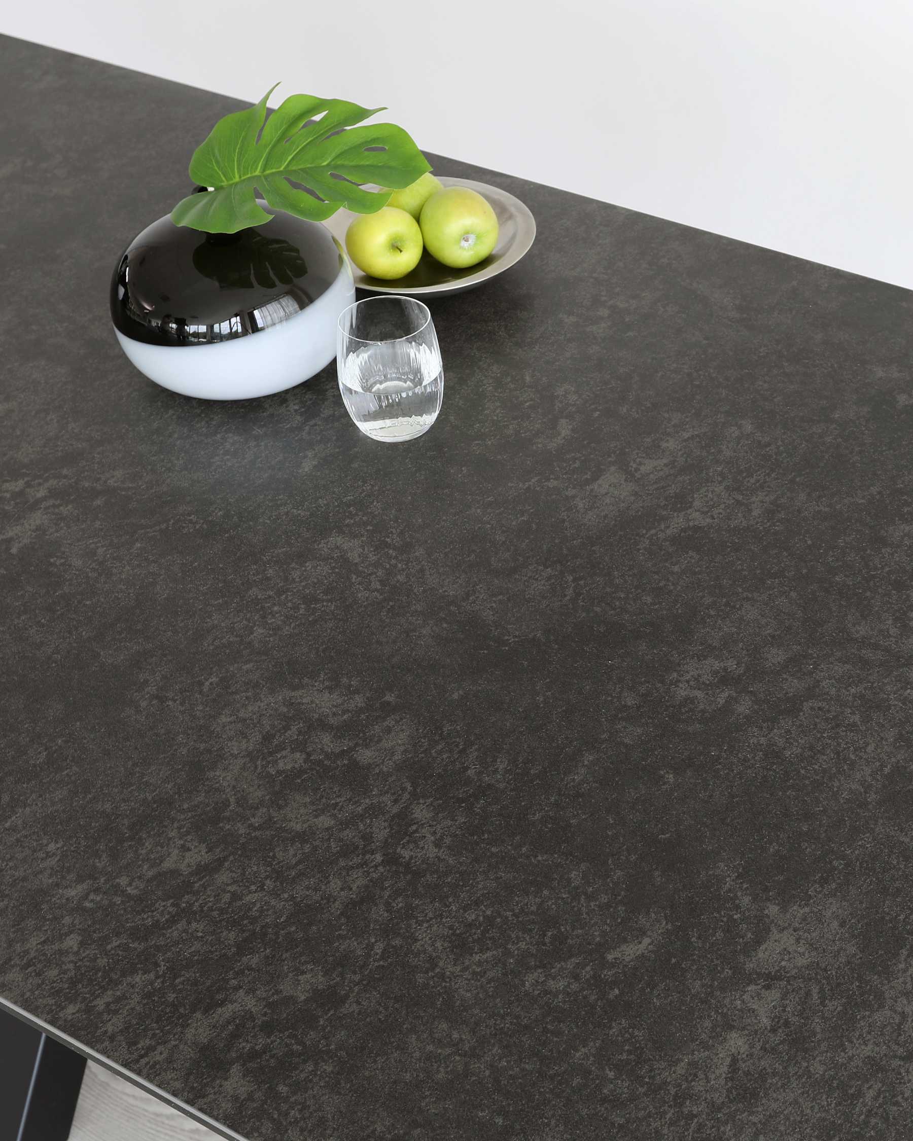 Modern minimalist dining table with a dark grey stone-textured top and thin metal legs, decorated with a small white and black round vase containing a Monstera leaf, a metallic oval tray with green apples, and a clear glass of water.