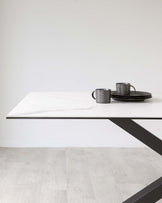 Modern minimalist dining table with a white marble top and angular black metal legs, accompanied by a set of two dark textured mugs and a coordinating black tray placed on top of the table, set against a neutral background with light wooden flooring.