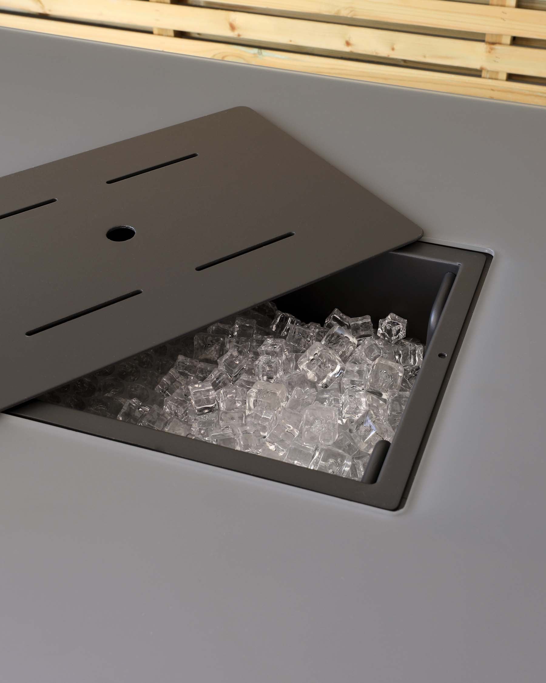 Close-up of a modern built-in ice storage bin with a grey sliding cover, partially open to reveal clear ice cubes inside. The bin appears to be integrated into a sleek grey countertop, suggesting a contemporary and functional design for commercial or high-end residential use.