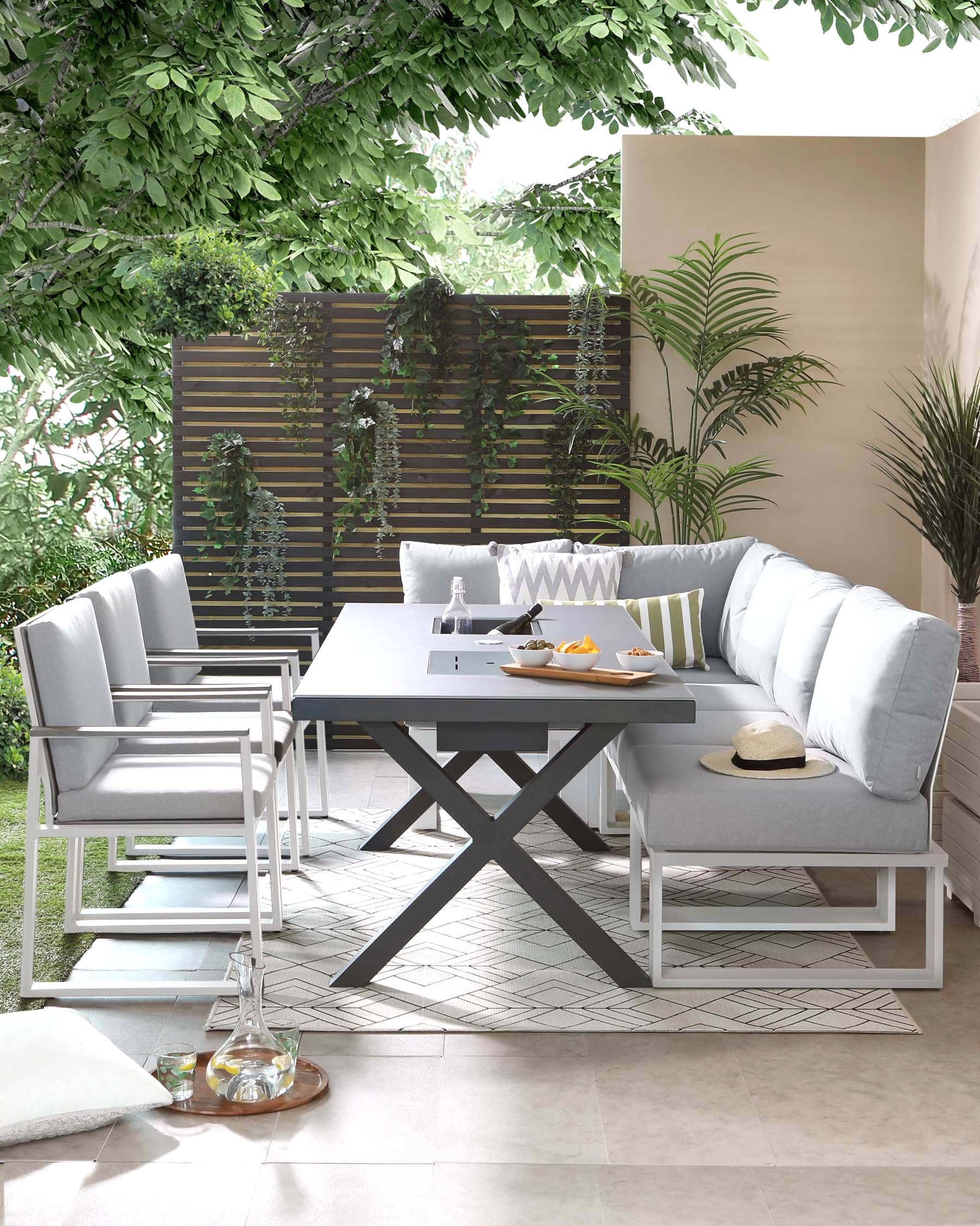 Outdoor dining set with a contemporary design, featuring a rectangular table with a grey finished top and white X-shaped legs, accompanied by two armchairs and a three-seater sofa with white metal frames and light grey cushions, arranged on a patio setting with greenery.