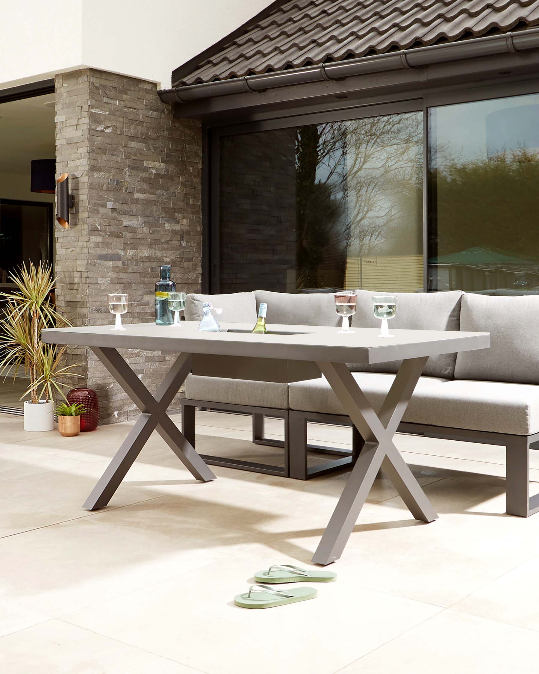 Modern outdoor dining furniture featuring a rectangular table with a solid top and bold X-shaped legs in a matte grey finish. Paired with a matching bench-style seat with plush light grey cushions for comfortable seating. The setting is complemented by a coordinating L-shaped corner lounge sofa, maintaining a seamless aesthetic. Ideal for sophisticated al fresco dining and relaxing.