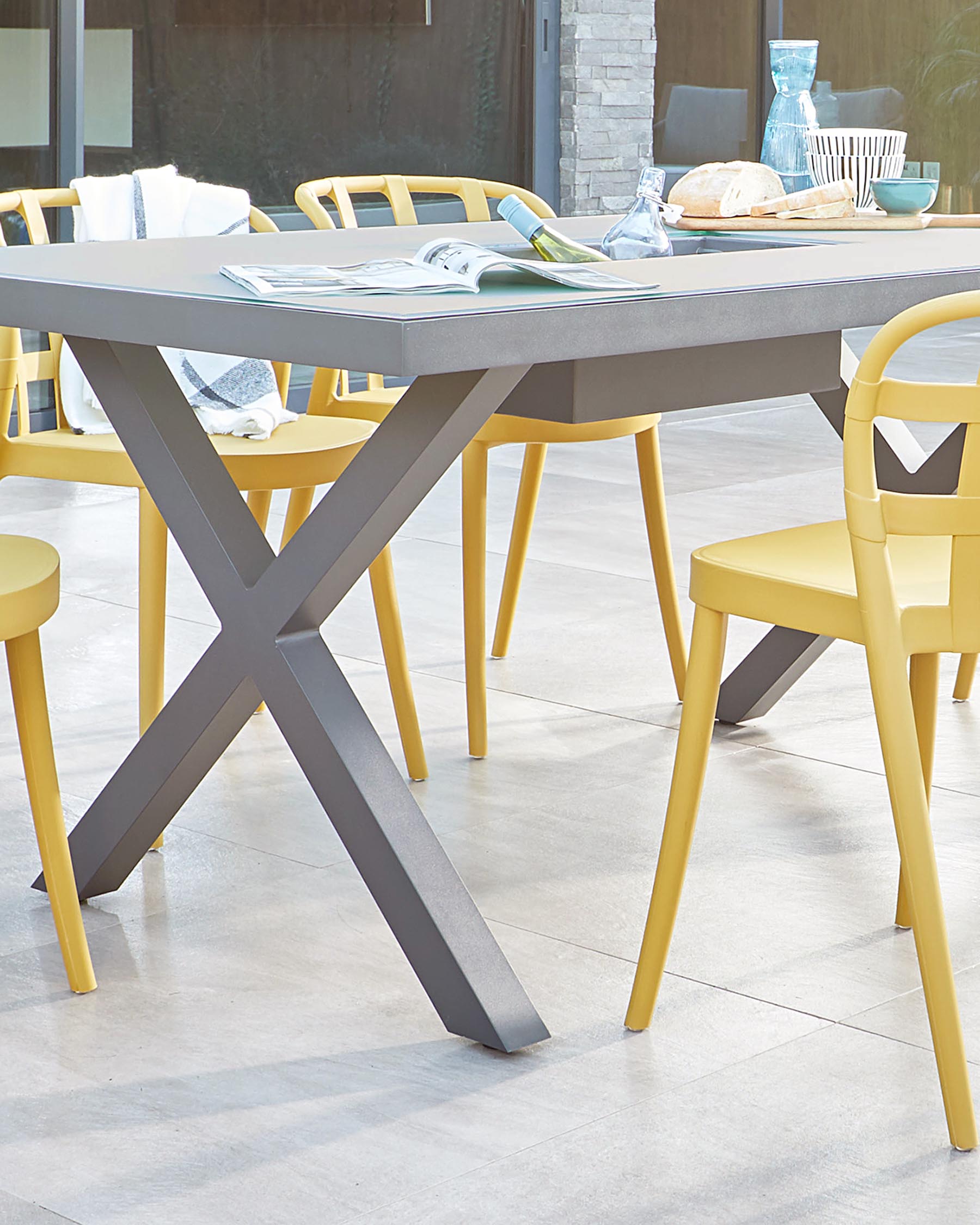 Modern outdoor dining furniture set featuring a stylish, rectangular, grey tabletop with a unique X-shaped base and complementing mustard yellow chairs with rounded backrests. The set is accessorized with a casual dining arrangement, hinting at effortless entertaining.