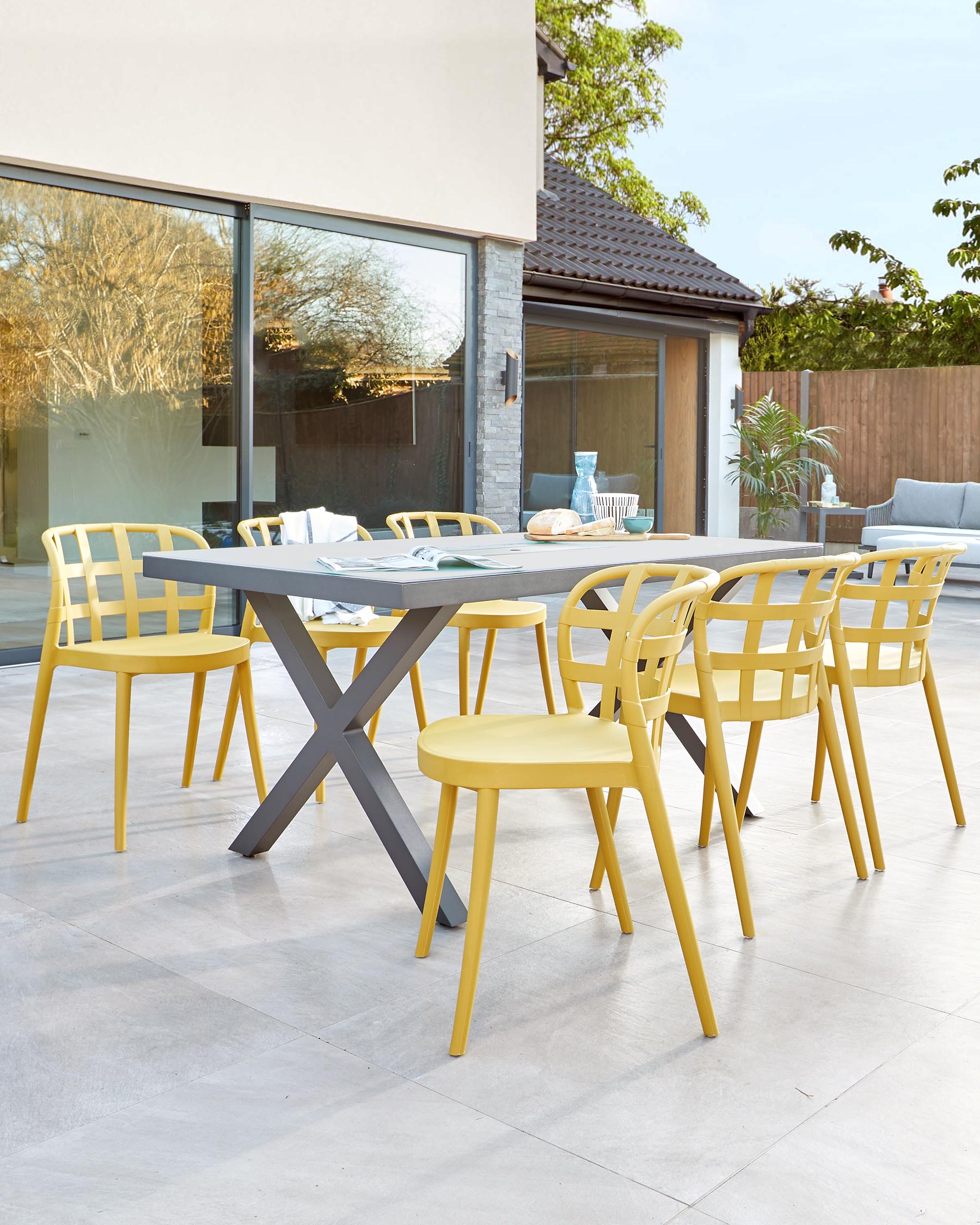 Modern outdoor dining furniture set featuring a rectangular table with a grey tabletop and crossed-leg design, paired with four vibrant yellow chairs with a geometric open-back pattern, displayed on a patio with a tiled floor adjacent to a large glass door leading to a house.