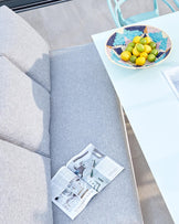 Modern outdoor patio set featuring a light grey, L-shaped sectional sofa and a sleek white coffee table with a decorative multi-coloured woven bowl containing lemons and limes on top.