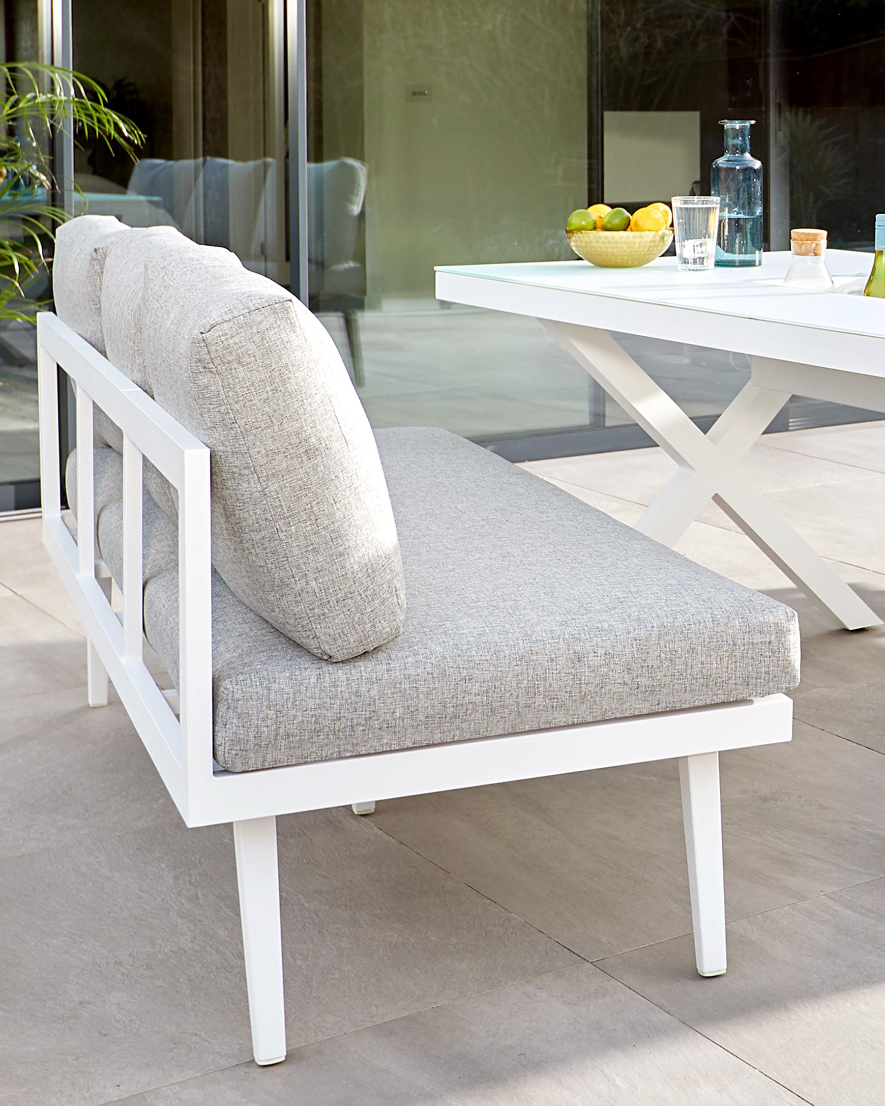 Modern outdoor lounge chair with a white frame and light grey cushions, accompanied by a white X-leg side table holding a glass jug, a bowl of lemons, and two glasses.