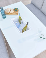 Modern white rectangular coffee table with a built-in ice bucket compartment, showcasing an innovative design for entertaining. The table features a sleek, minimalist style with a glossy finish, set against an outdoor patio setting with a grey sofa. On the table, there's a wooden serving tray with bread and olives, alongside a bottle of wine, a water bottle on ice, a drinking glass, and a pair of sunglasses, emphasizing a relaxed, leisurely atmosphere.