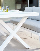 Modern outdoor furniture featuring a white minimalist dining table with a sleek tabletop and crossed leg design, paired with a coordinating bench that has a light grey cushion. The set suggests a contemporary, clean aesthetic suitable for a patio or terrace setting.