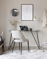 Modern minimalist home office with a sleek black metal console table featuring a glass top and slender angled legs, paired with a light grey upholstered chair with a curved backrest and slim black metal legs. The chair is positioned to suggest use as a desk chair for the console table workspace.