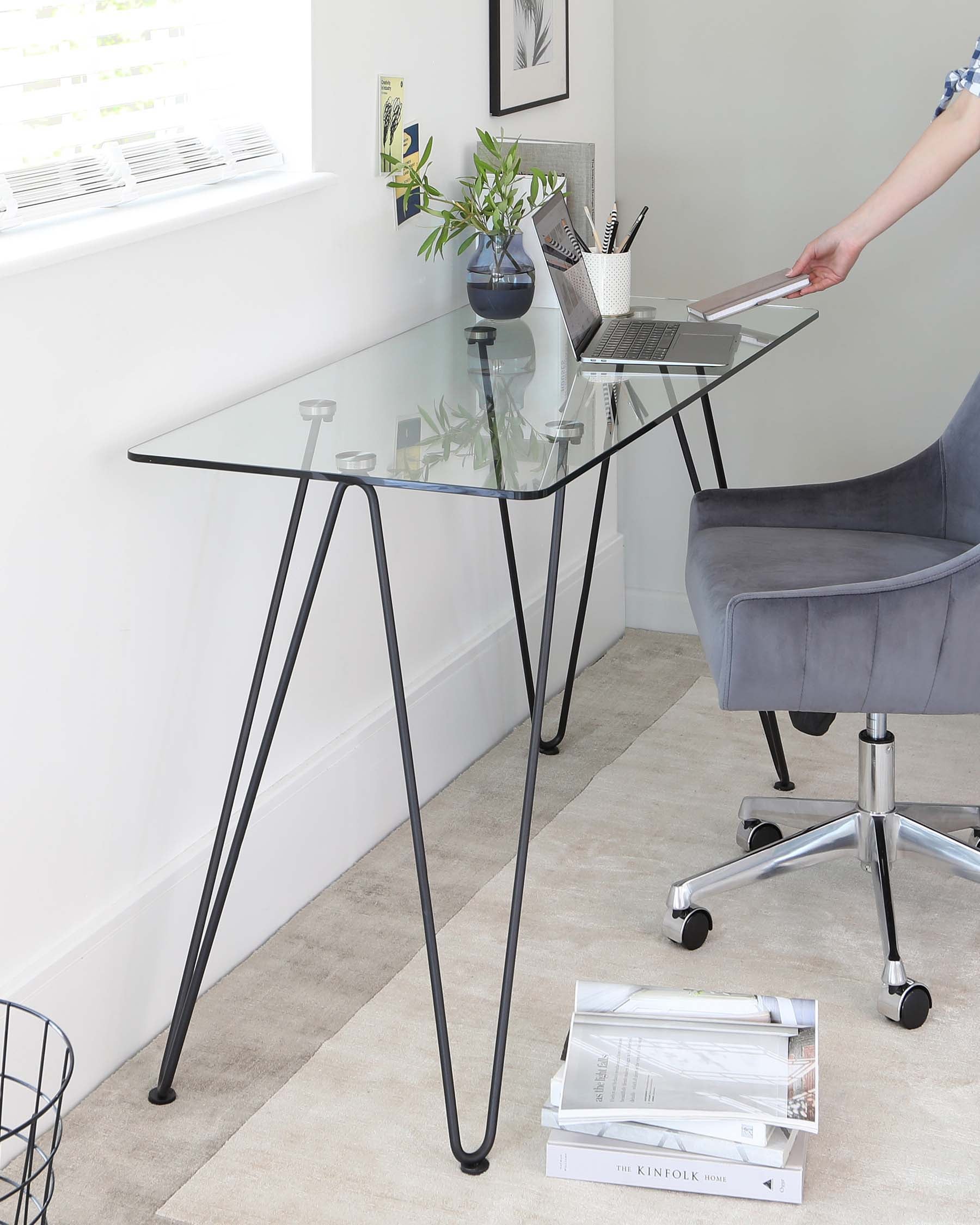 Modern minimalist glass top desk with slender black metal legs and adjacent grey fabric swivel office chair on casters.