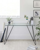 A minimalist modern desk with a clear glass tabletop and sleek black metal hairpin legs.