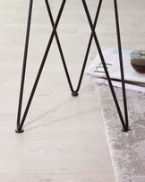 A close-up of a modern minimalist table leg design with a crossed steel frame in black, standing on a pale hardwood floor with a part of a grey area rug visible.