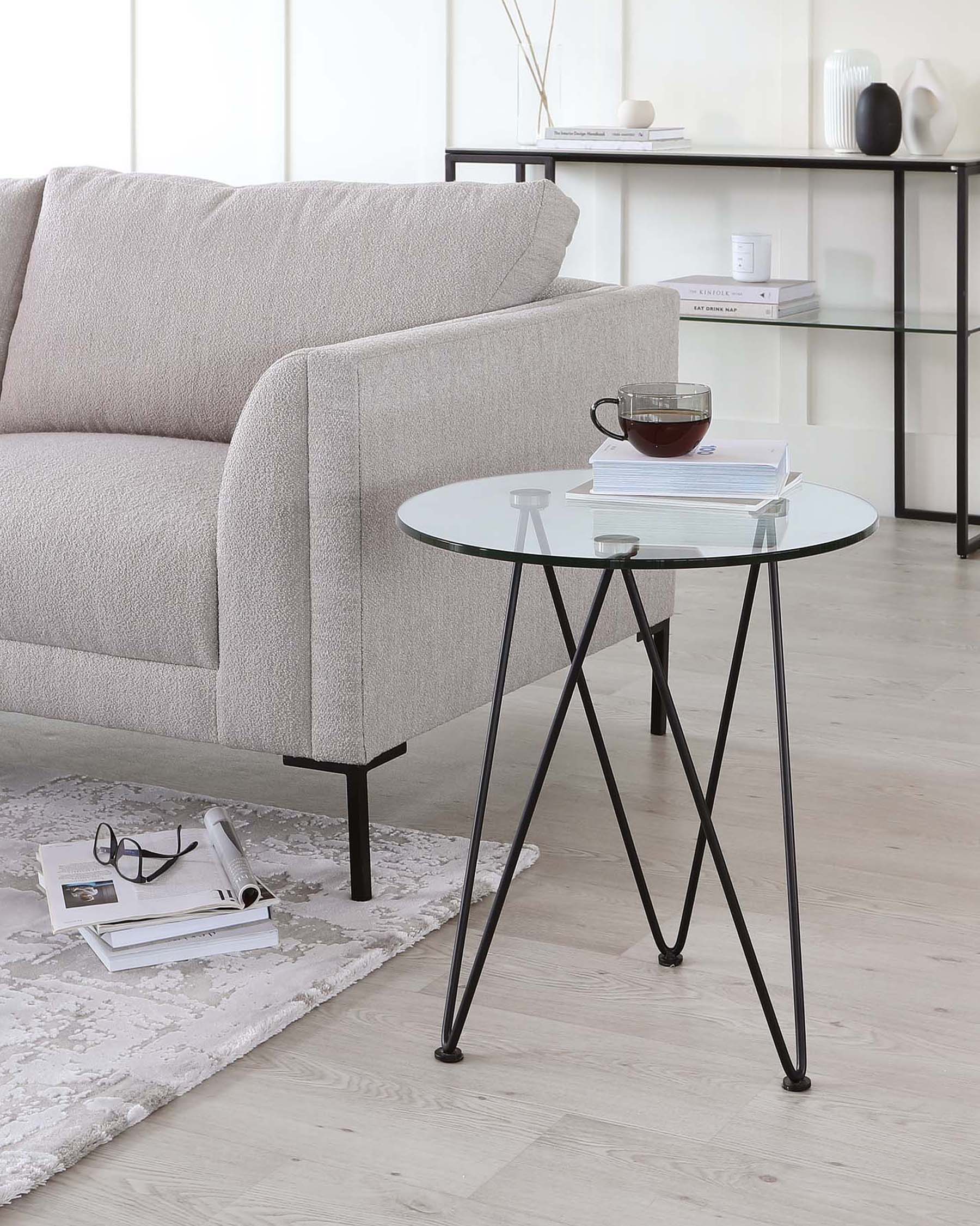 A modern, minimalist living room setup featuring a light grey textured fabric sofa with clean lines and a rounded armrest. Adjacent to the sofa is a round, glass-topped side table with a thin black metal frame forming a tripod support. On the table is a simple ceramic mug and a small stack of books. The setting includes a patterned off-white area rug, a sleek black metal shelving unit in the background holding decorative items, and a second stack of books and glasses on the floor beside the side table.