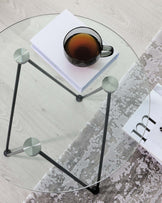 A modern round glass-topped coffee table with a minimalist black metal frame. The table features three sleek, rod-like legs connected by an equilateral triangular metal base. Each leg is capped with a disc at the top where it meets the glass surface, adding a subtle detail to the design. The table is positioned on a textured grey and white area rug over a light wood flooring. Resting on the glass surface are a white book and a black ceramic cup filled with a dark liquid.