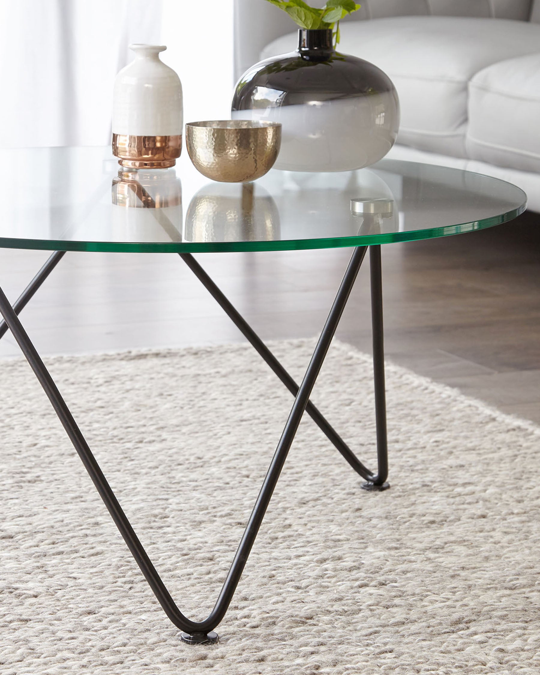 Round glass-top coffee table with intersecting black metal legs on a textured cream rug.