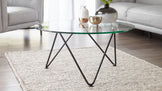 Modern round glass-top coffee table with black metal legs arranged in a crisscross pattern, placed on a textured off-white area rug with a section of a white leather sofa in the background.