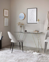 Elegant, modern furniture including a sleek glass-top desk with slender gold legs and black feet, paired with a light grey upholstered chair with a curved backrest and black legs.