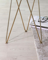 Sleek golden metal legs in a crossed design supporting a minimalist table, on a pale wood floor next to a soft textured off-white area rug with a magazine and eyeglasses resting on top.