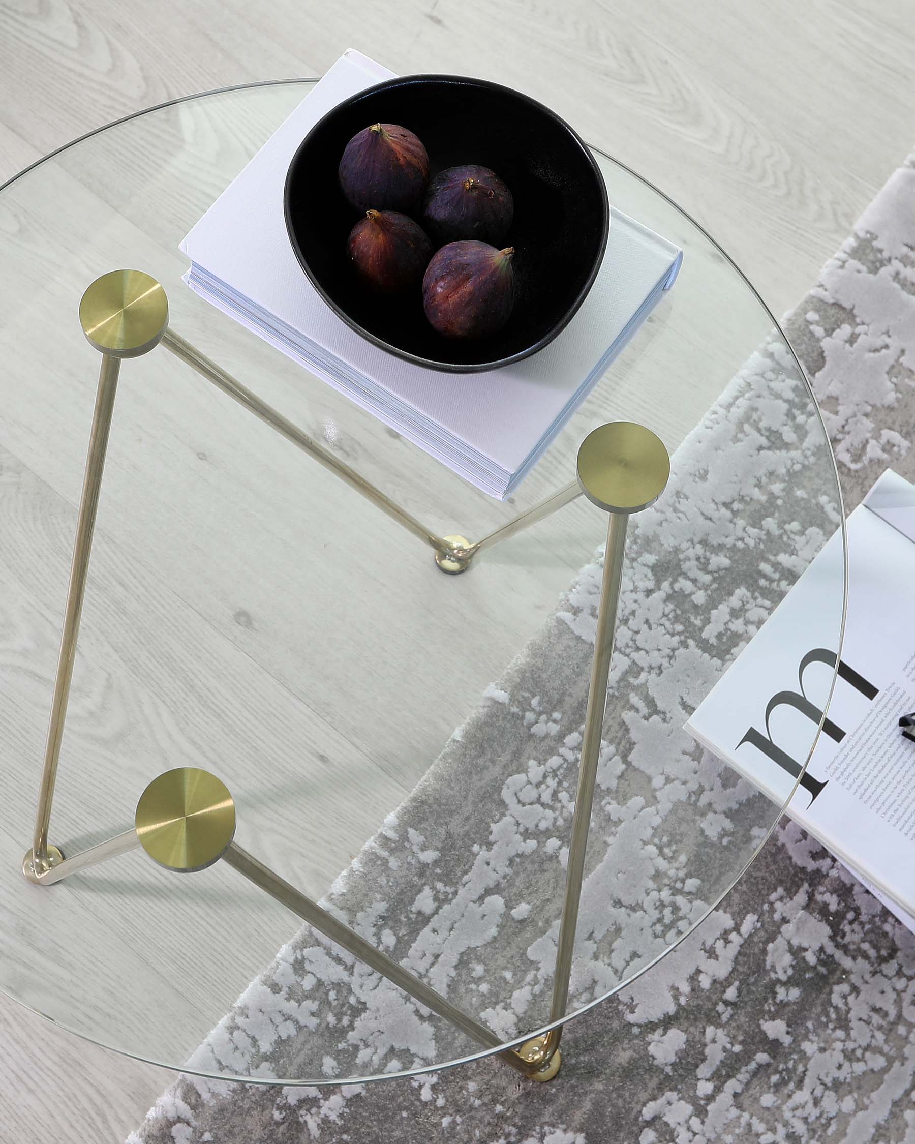 Round glass coffee table with a sleek brass frame, featuring three legs with ballpoint accents, positioned on a grey and white patterned area rug. On the table's surface, there's a bowl with fruit on top of a stacked set of white books next to a magazine.