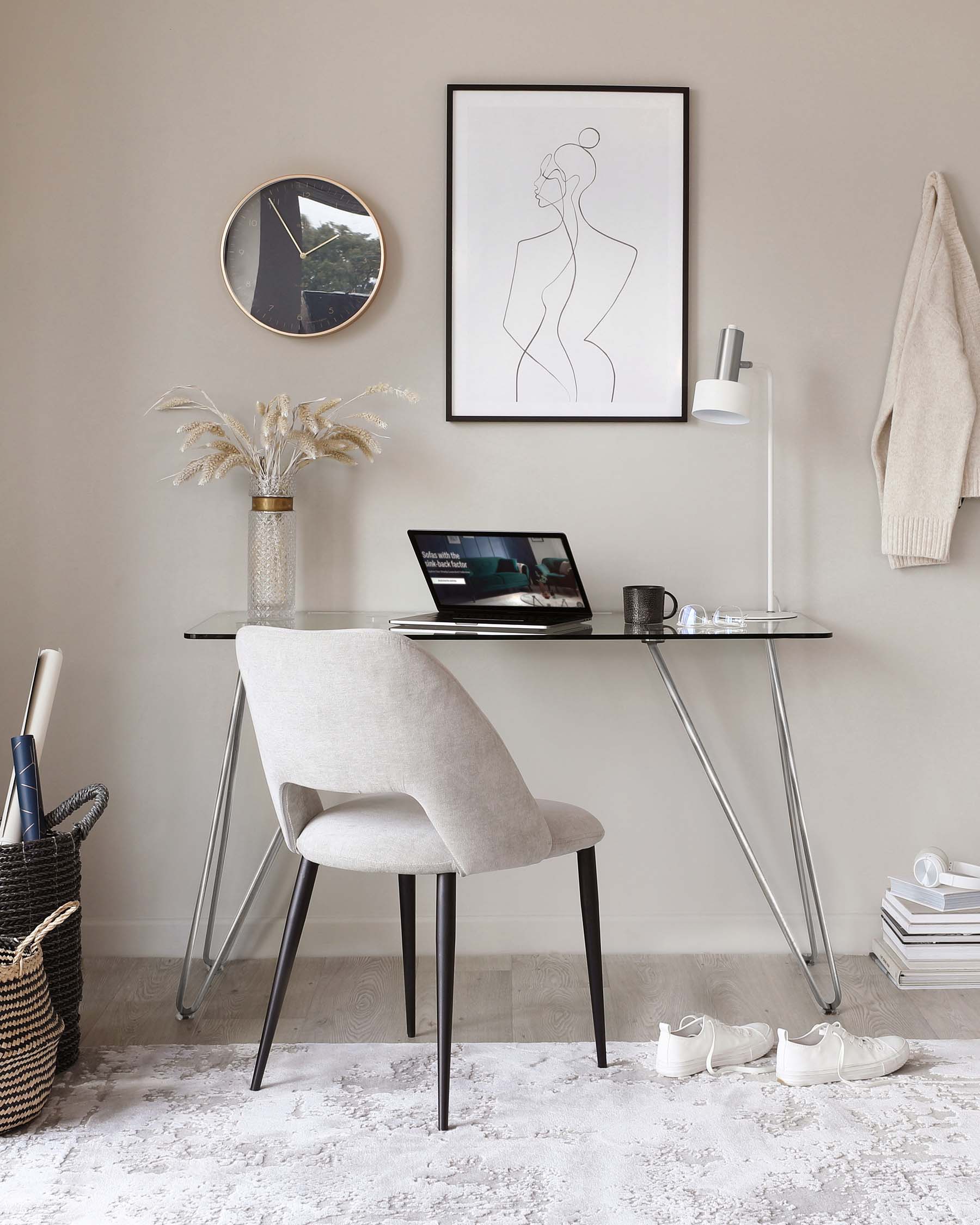 Modern minimalist home office setup featuring a sleek glass-top desk with hairpin metal legs and a comfortable upholstered chair with a curved backrest and black legs. The setup is complemented by a textured area rug underfoot.