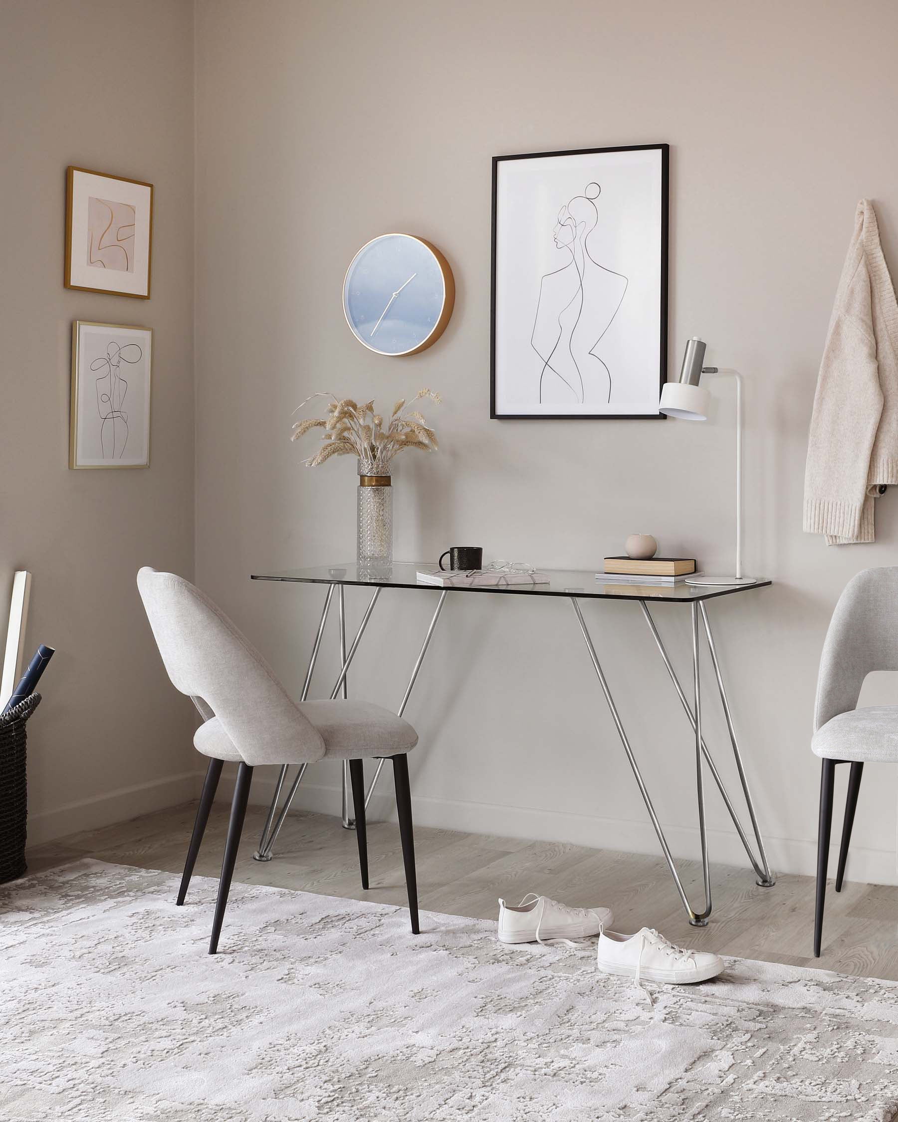 Modern minimalist home office setup featuring a sleek glass-top desk with slender metal legs. Paired with the desk are two upholstered chairs in light fabric with cushioned seats and backs, mounted on slim black wooden legs. The scene is situated on a textured white area rug.