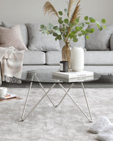 A contemporary light grey fabric sofa with plush cushions and a textured throw. In front, a round black coffee table with a unique X-shaped metallic base. The table is styled with decorative books and a modern white vase with greenery. The room features a soft grey rug with subtle texture.