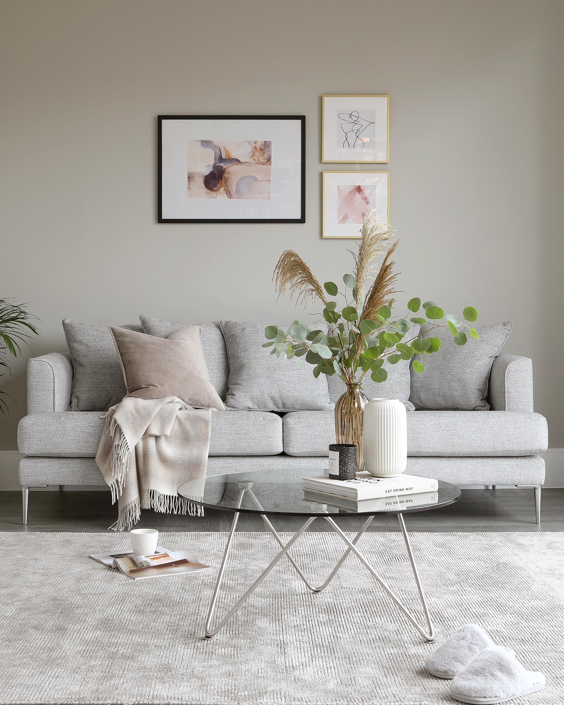 Three-seat grey fabric sofa with linear design and multiple throw pillows in shades of grey and taupe, accompanied by a lightweight round white coffee table with metal legs. A neutral-toned woven throw blanket drapes over one side of the sofa. A textured grey area rug underlines the furniture set-up.