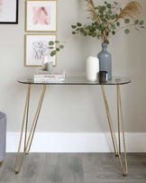 An elegant modern console table with a sleek, smoked glass top and slender, gently tapered, golden metal legs. The table is adorned with a curated selection of decorative items including books, a striated white vase, a muted blue bottle vase, a speckled black tumbler, and a small plant, and is positioned against a neutral wall with framed abstract art pieces above.