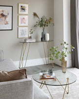 Elegant contemporary living room with two pieces of furniture. On the left is a slender console table with a dark top and golden metal legs in an airy, intersecting design. On the right is a round coffee table with a clear glass top and matching golden metal legs that taper to a point, mirroring the aesthetic of the console table. Both pieces reflect a modern minimalist style and compliment the neutral colour palette of the space.