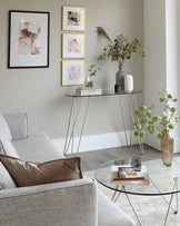 Modern living room with a minimalist grey fabric sofa, a round glass coffee table with black metal legs, and a narrow console table with a black frame and glass top, accessorized with vases and books.