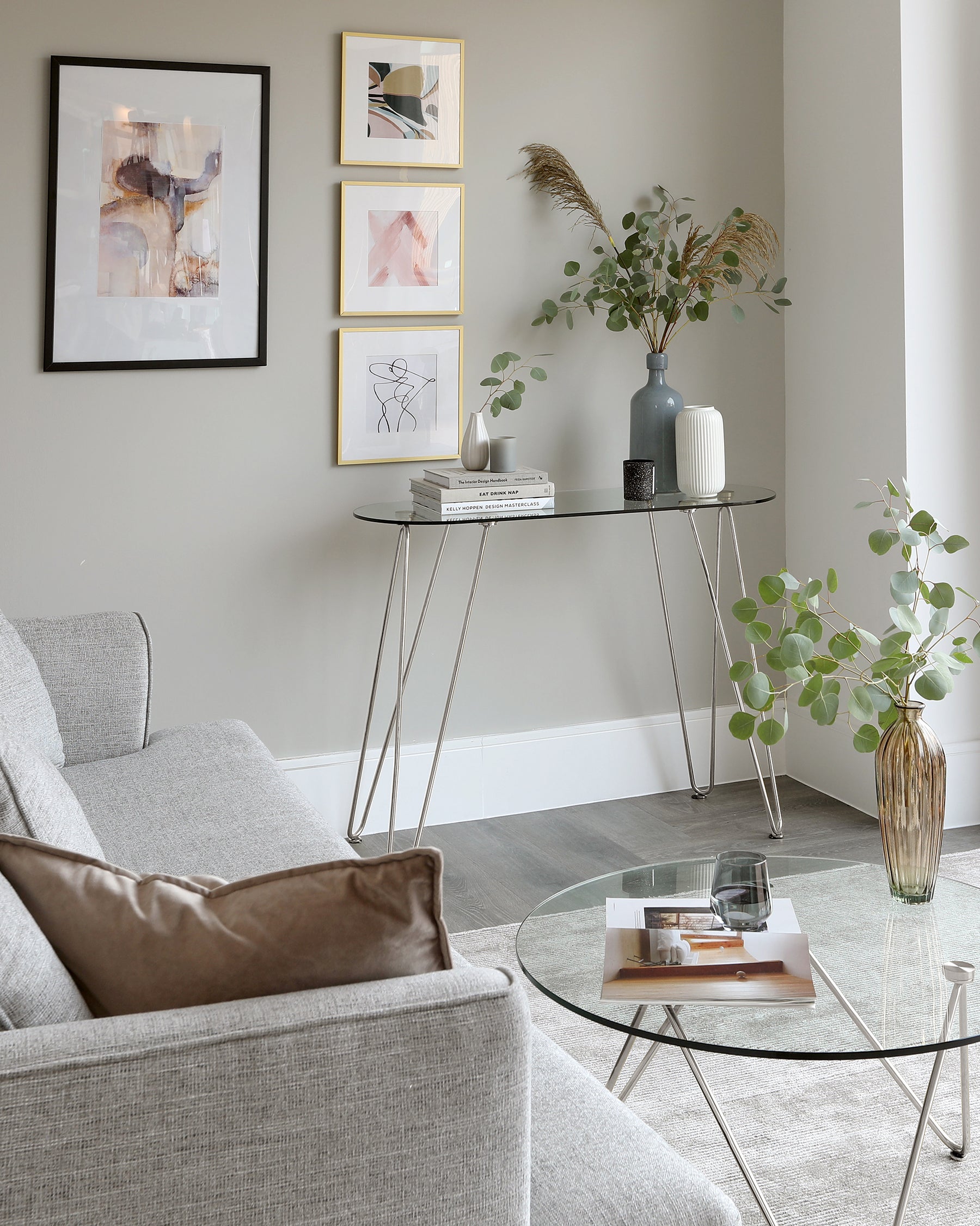 Modern living room with a minimalist grey fabric sofa, a round glass coffee table with black metal legs, and a narrow console table with a black frame and glass top, accessorized with vases and books.