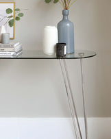 A modern semi-circular console table with a sleek glass top and metallic, slender, crisscrossed legs.