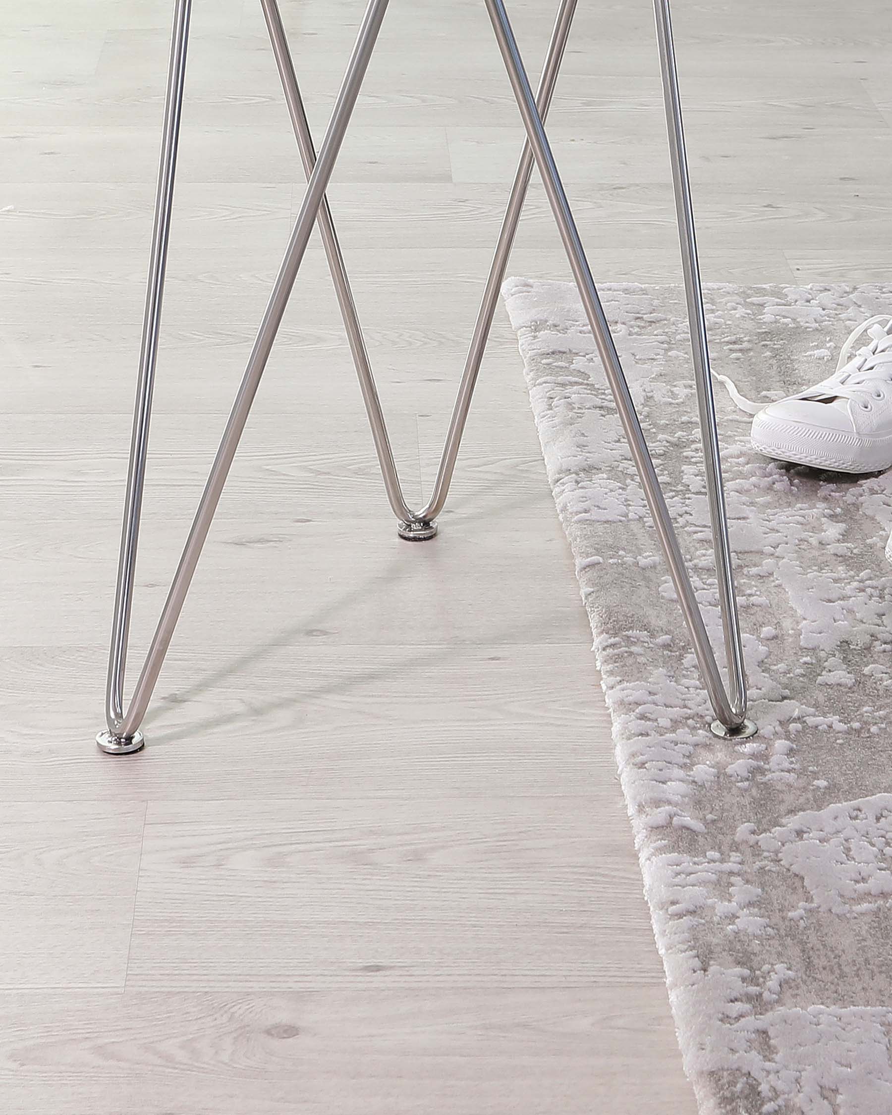 Modern minimalist chair with sleek chrome legs and visible portion of a light-coloured textured rug on a wooden floor; the edge of a white sneaker can also be seen.