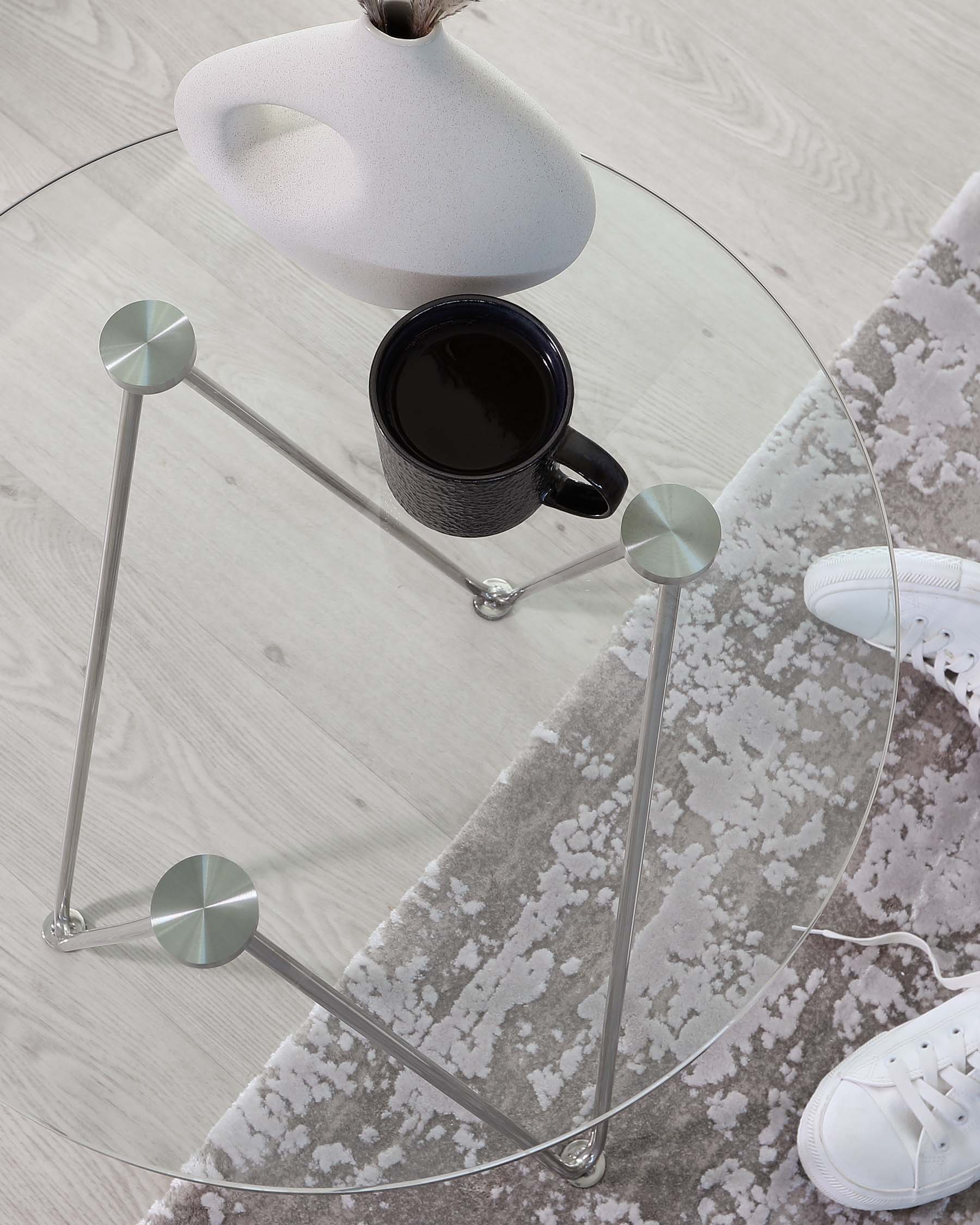 A contemporary round glass-top side table with a minimalistic chrome frame and legs, featuring disc-shaped feet. On its surface rests a sleek black coffee mug and a modern ceramic vase.