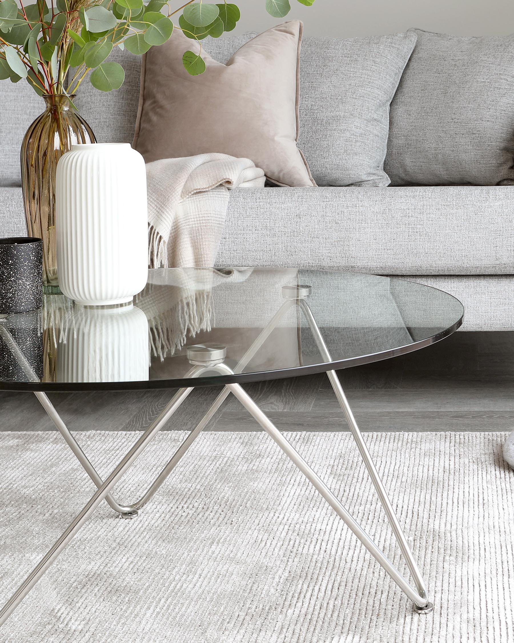 Elegant living room setting featuring a modern, heathered grey fabric sofa with plush cushions and a neutral-toned throw blanket. In the foreground, a sleek, round glass coffee table with a unique, tripod-style silver metallic base is visible, resting on a textured ivory area rug.