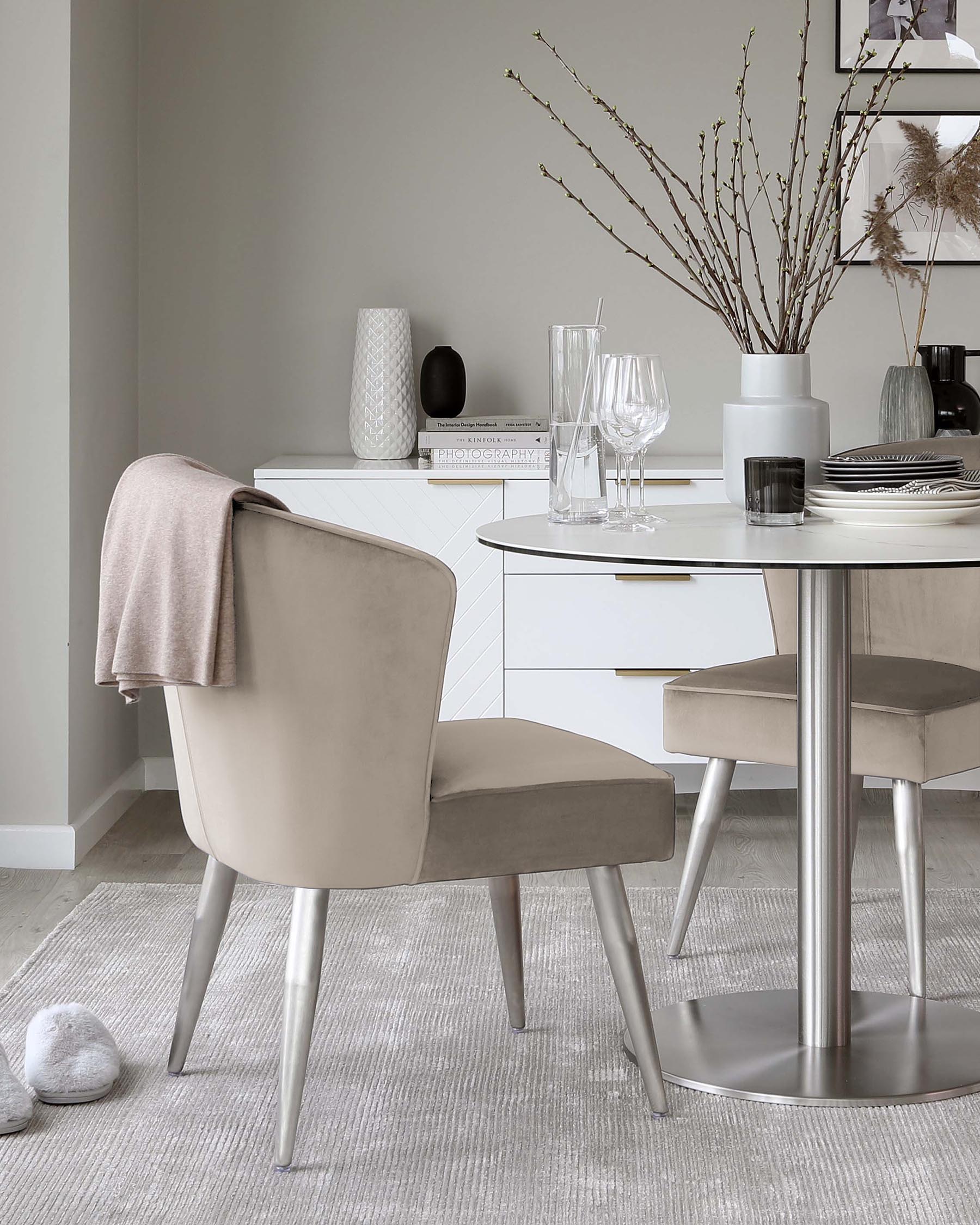 Modern dining room furniture featuring a sleek white round table with a reflective stainless steel base and a pair of elegant taupe upholstered chairs with slender metal legs.