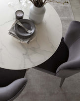 A modern, circular dining table with a white marble top and a dark central base, accompanied by elegant, grey upholstered dining chairs with slender legs. The table is set with striped monochrome plates, silverware, and a vase with branches, creating a sophisticated dining arrangement.