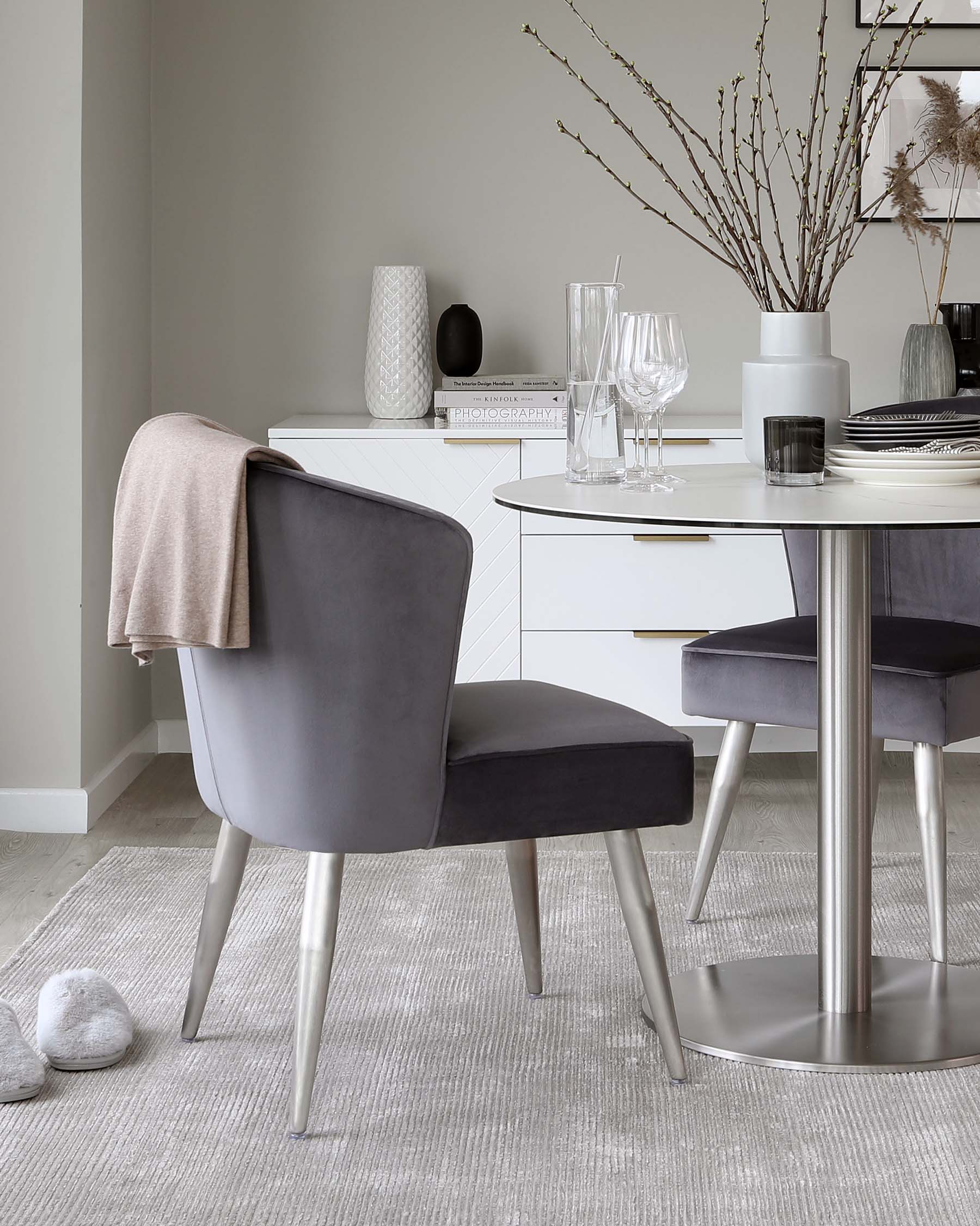 Modern dining room with a round white table with a silver metallic base, paired with elegant grey upholstered chairs with metal legs, set on a textured light grey area rug. A white sideboard adorned with decorative vases, books, and dishware complements the minimalist space.