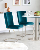 Two modern velvet dining chairs in a rich teal colour featuring sleek metal cantilever bases, paired with a round white-top dining table with a metal base.