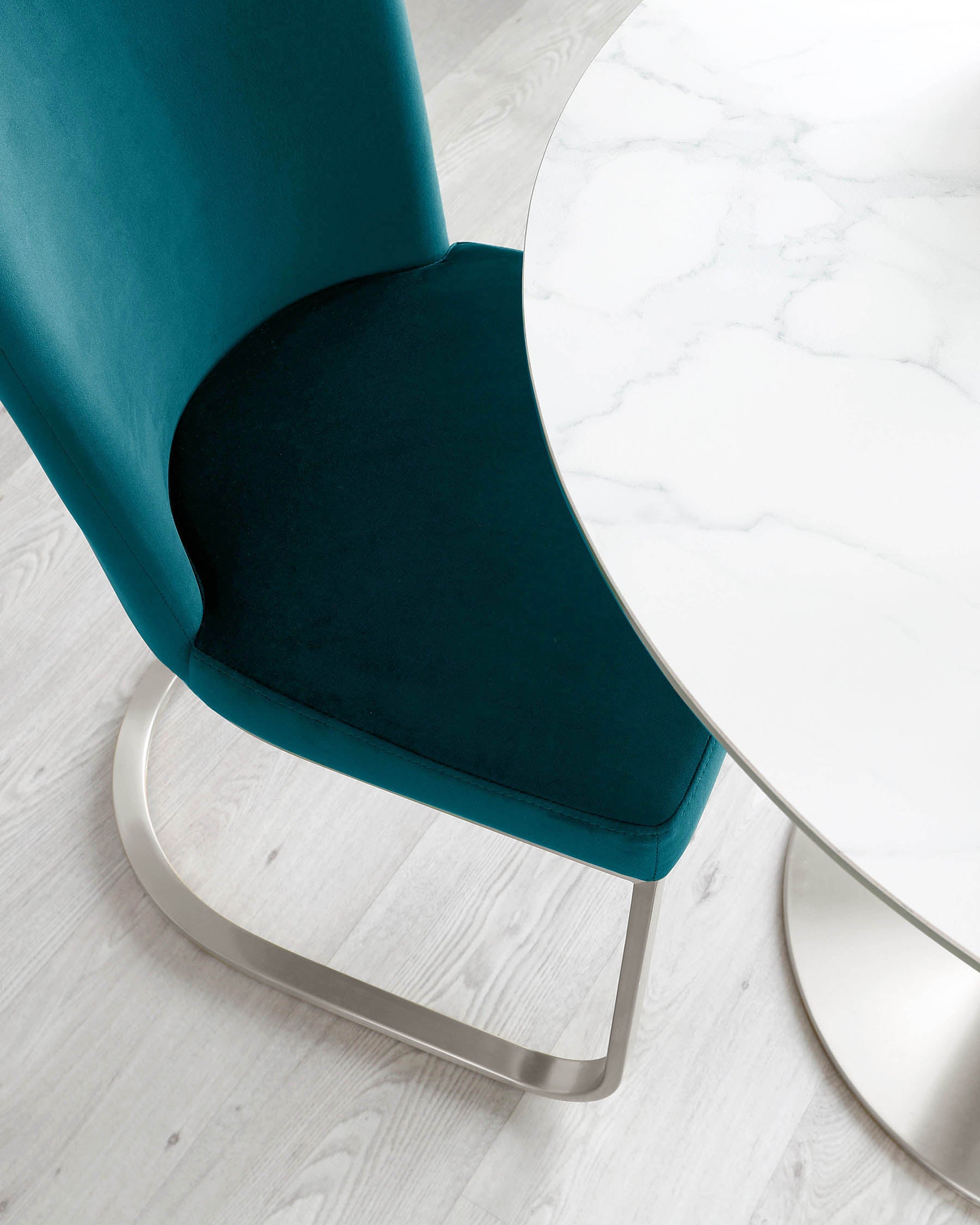 Modern teal blue velvet chair with a smooth, curved backrest and a silver metal base alongside a round marble top table with a reflective metal base, displayed on a light wood laminate flooring.