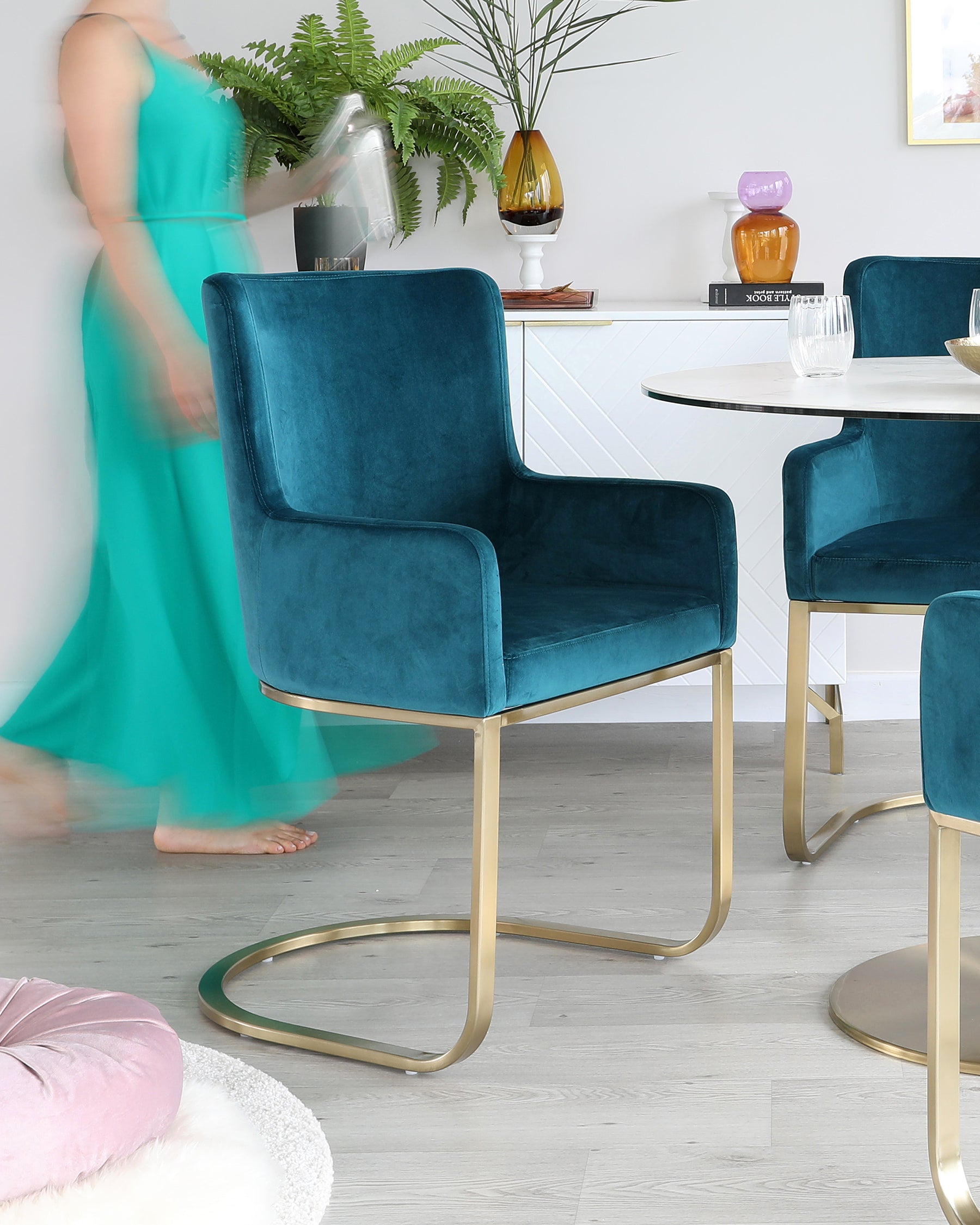 Velvet upholstered dining chairs in rich teal with smooth, curved backrests and luxurious gold-finished metal legs that wrap around the frame, paired with a sleek white round dining table featuring a complementary gold-finished metal base.