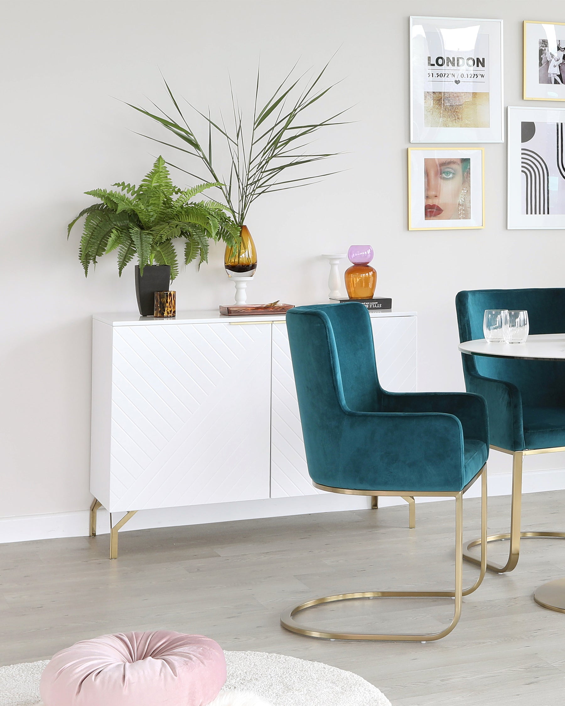 Modern furniture set in a stylish living room that includes a white geometric-patterned sideboard with gold metal legs, two luxurious teal velvet armchairs with curved gold metal frames, and a round blush floor cushion.