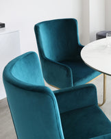 Elegant velvet dining chairs in a lush teal colour with a smooth curved back and armrests, paired with a sleek white marble tabletop with gold accents on the legs, showcased in a modern interior setting.