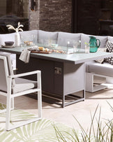 Contemporary outdoor furniture set featuring a sleek rectangular dining table with a frosted glass top and integrated cabinet storage, paired with a modern aluminium frame chair with light grey cushioned seat and backrest. The ensemble is complemented by a matching bench-style seat with comfortable back cushions adorned with patterned throw pillows.